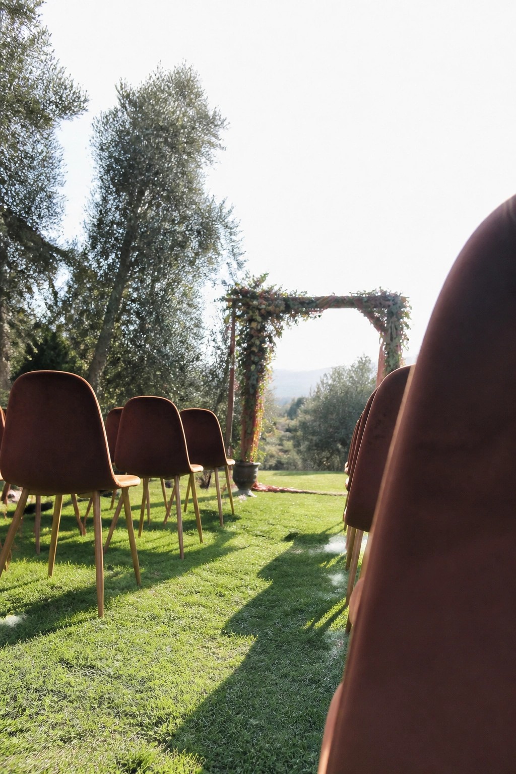 Rows of terracotta velvet chairs on green grass face a flower covered wooden arch in a sunny outdoor meadow with trees and hills beyond.