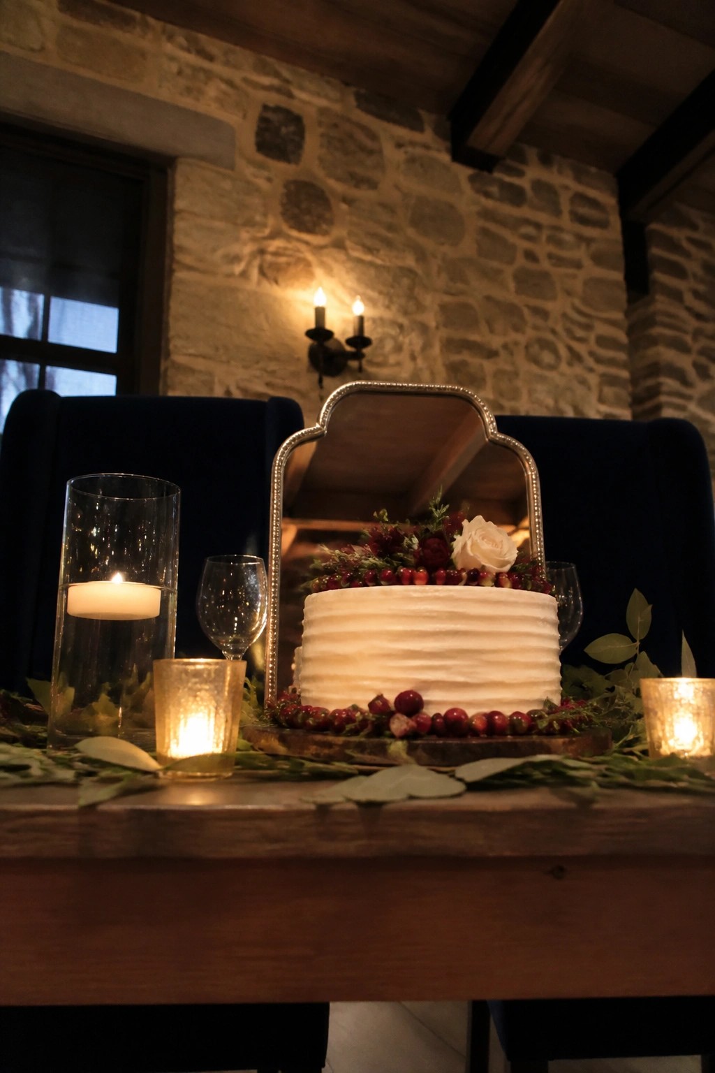 A white wedding cake with red cranberries and white roses is displayed on a wooden table with candles and greenery in front of navy chairs and a stone wall.