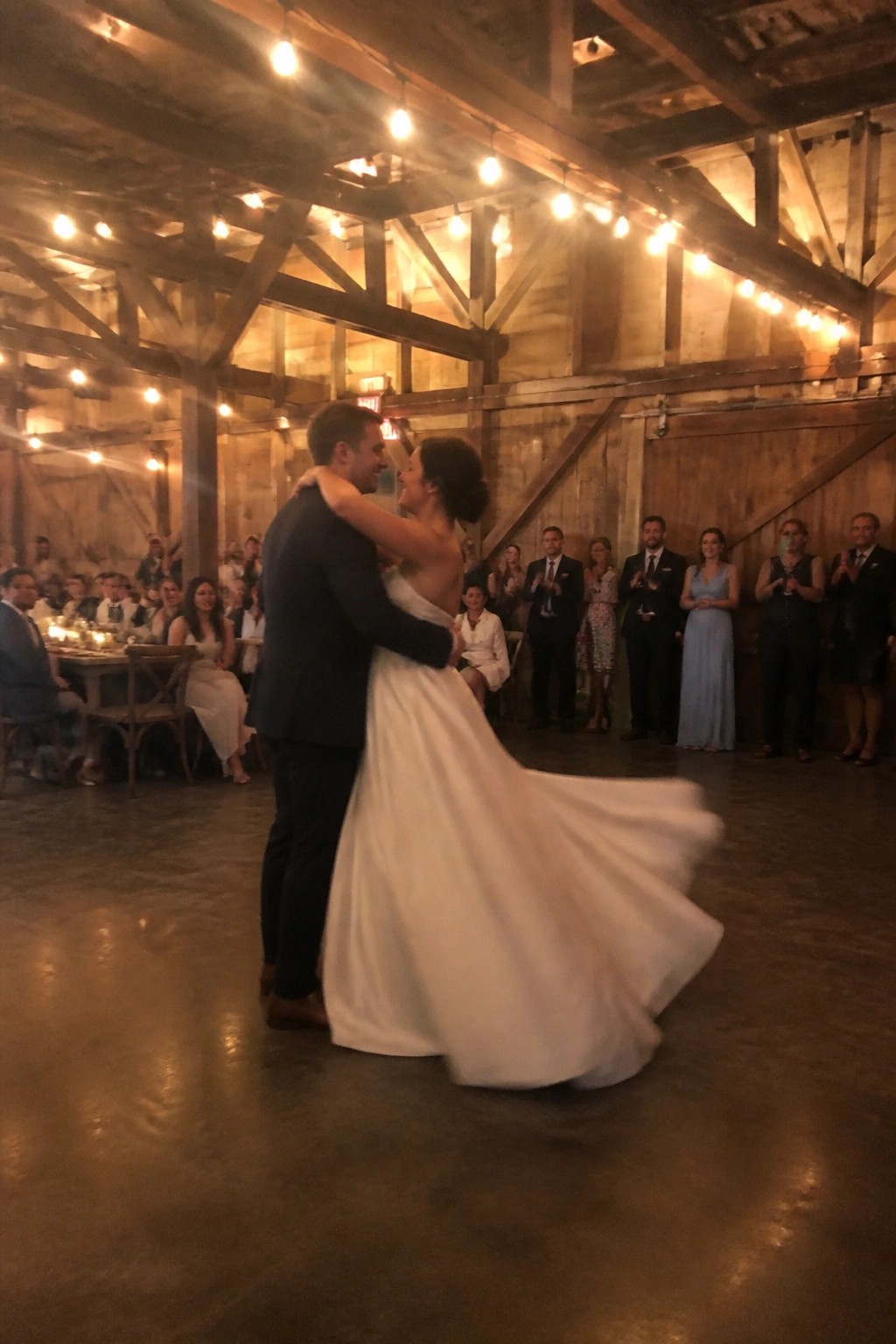 A bride in a flowing white dress and groom in a dark suit dance closely in a rustic barn lit by warm string lights as guests watch and applaud.