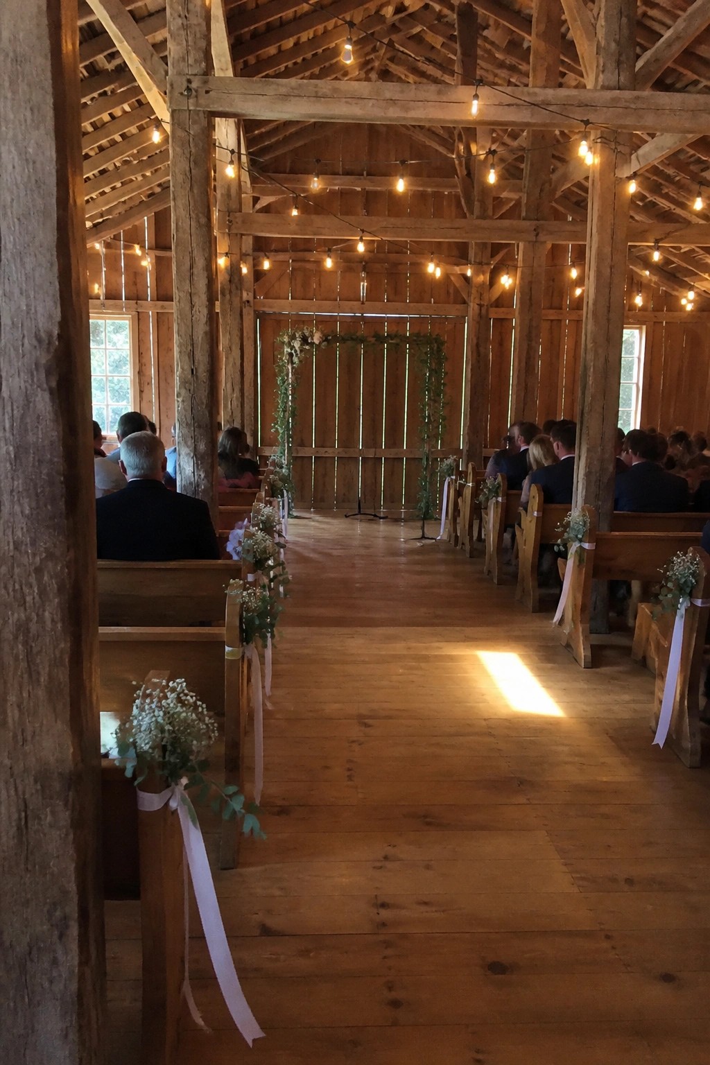 Interior of a rustic barn wedding venue with wooden pews decorated in white ribbons and flowers, string lights overhead, and an aisle leading to a greenery arch.