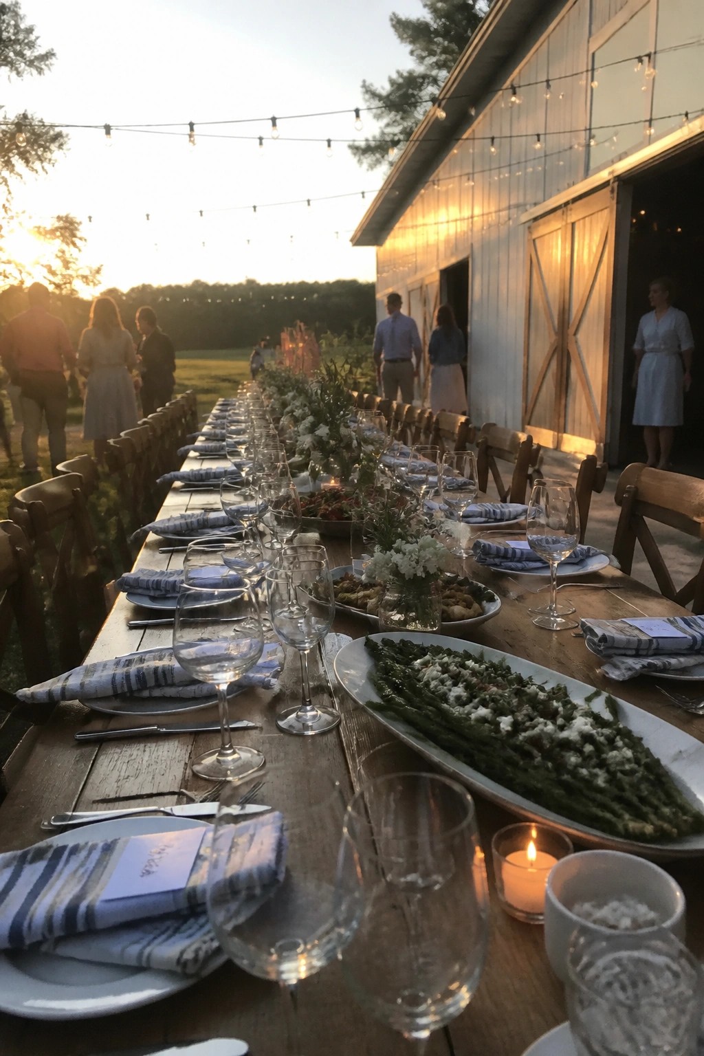 Long rustic wedding table set with blue napkins and food platters beside a white barn at sunset with string lights overhead.