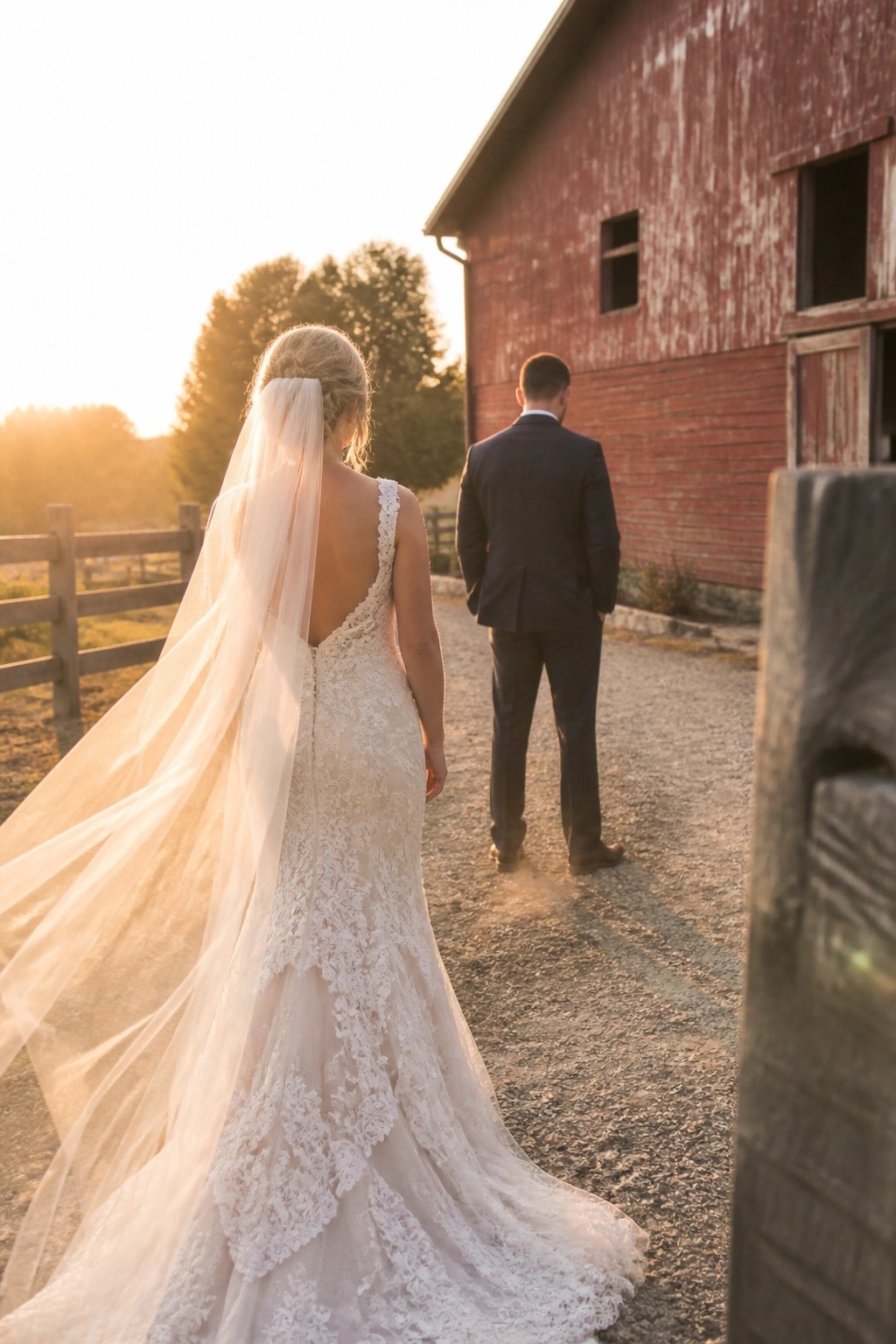 Bride in lace dress and veil follows groom in suit on gravel path beside red barn during golden hour sunset