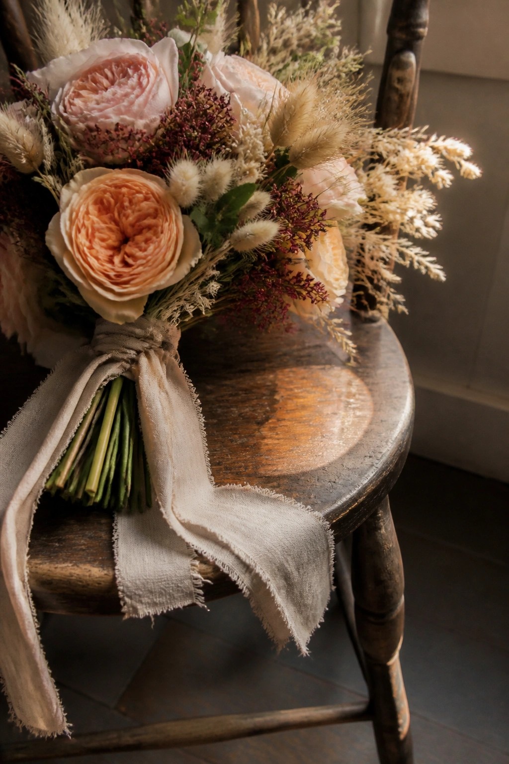 A rustic wedding bouquet of peach and pink roses with dried grasses and beige ribbon rests on a dark wooden chair in soft natural light.