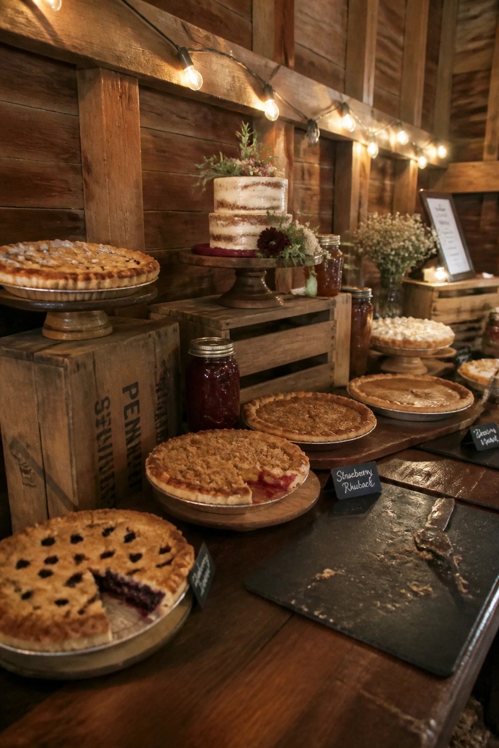 Rustic barn wedding dessert table with assorted pies a two tier cake string lights wooden crates and warm lighting.