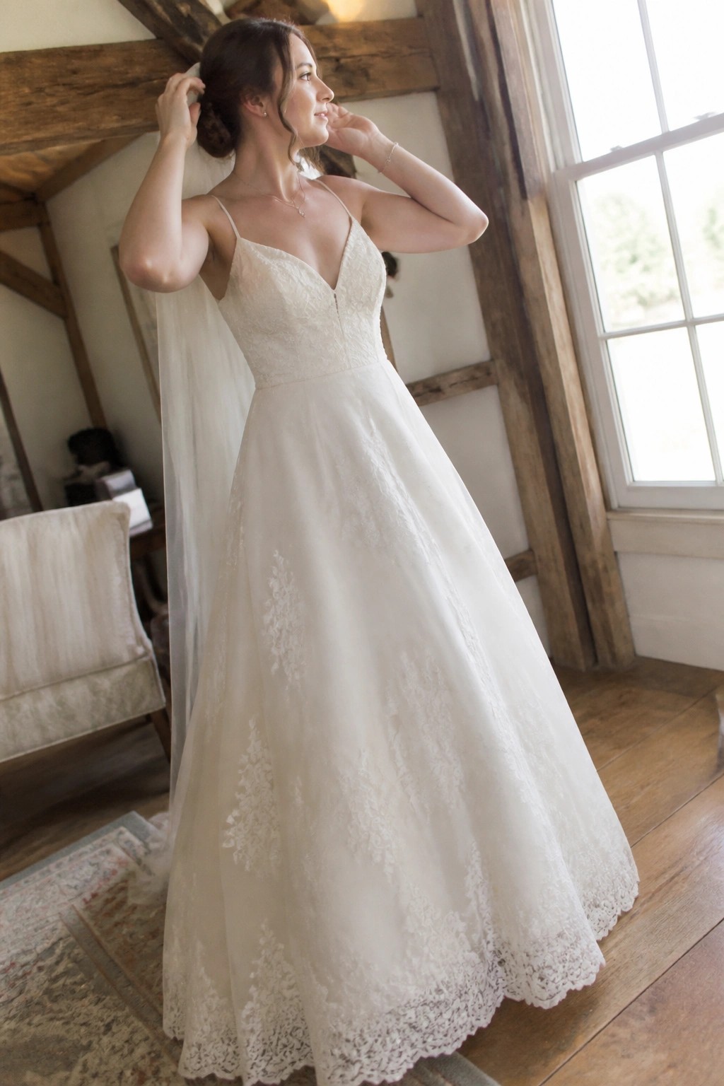 Bride in lace wedding dress with veil posing in rustic barn with wooden beams and window light