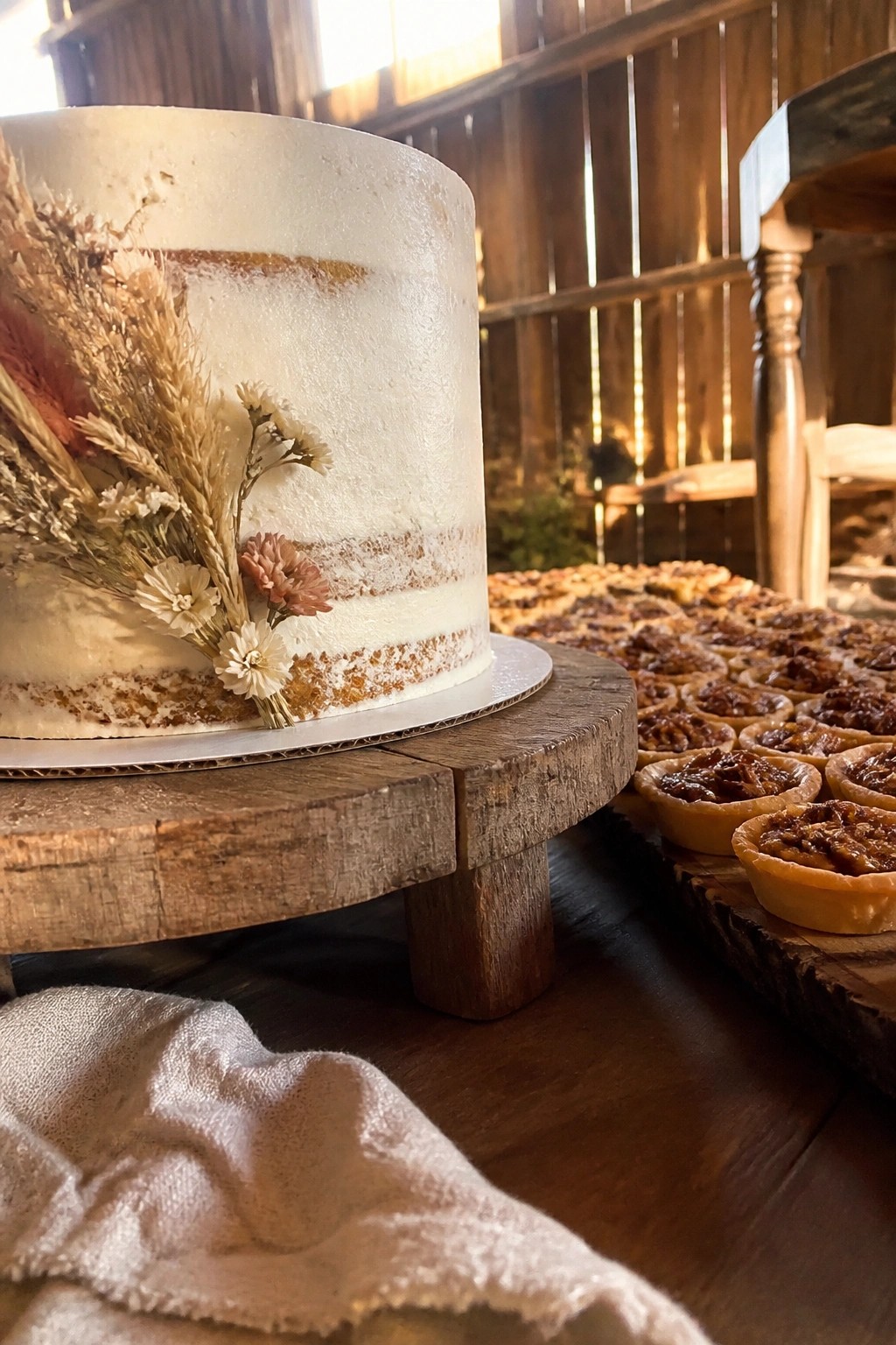 White wedding cake with dried flowers on wooden stand beside mini pecan tarts in rustic barn setting