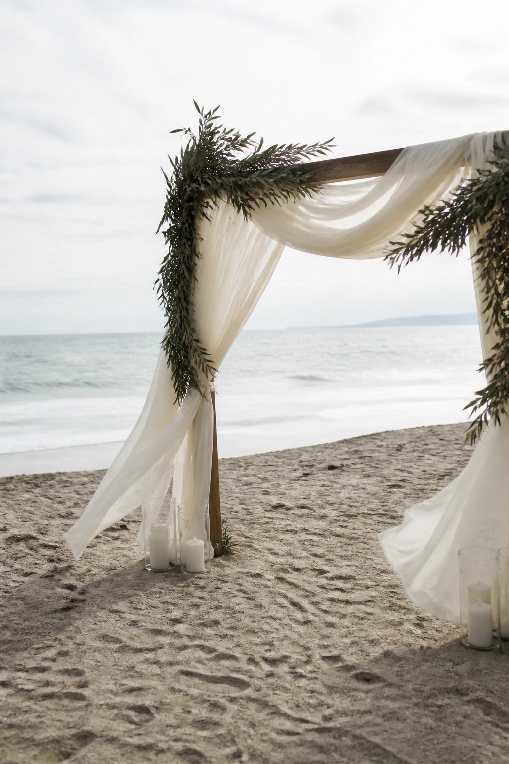 Beach wedding arch with white fabric drapes green foliage and candle holders on the sand facing the ocean