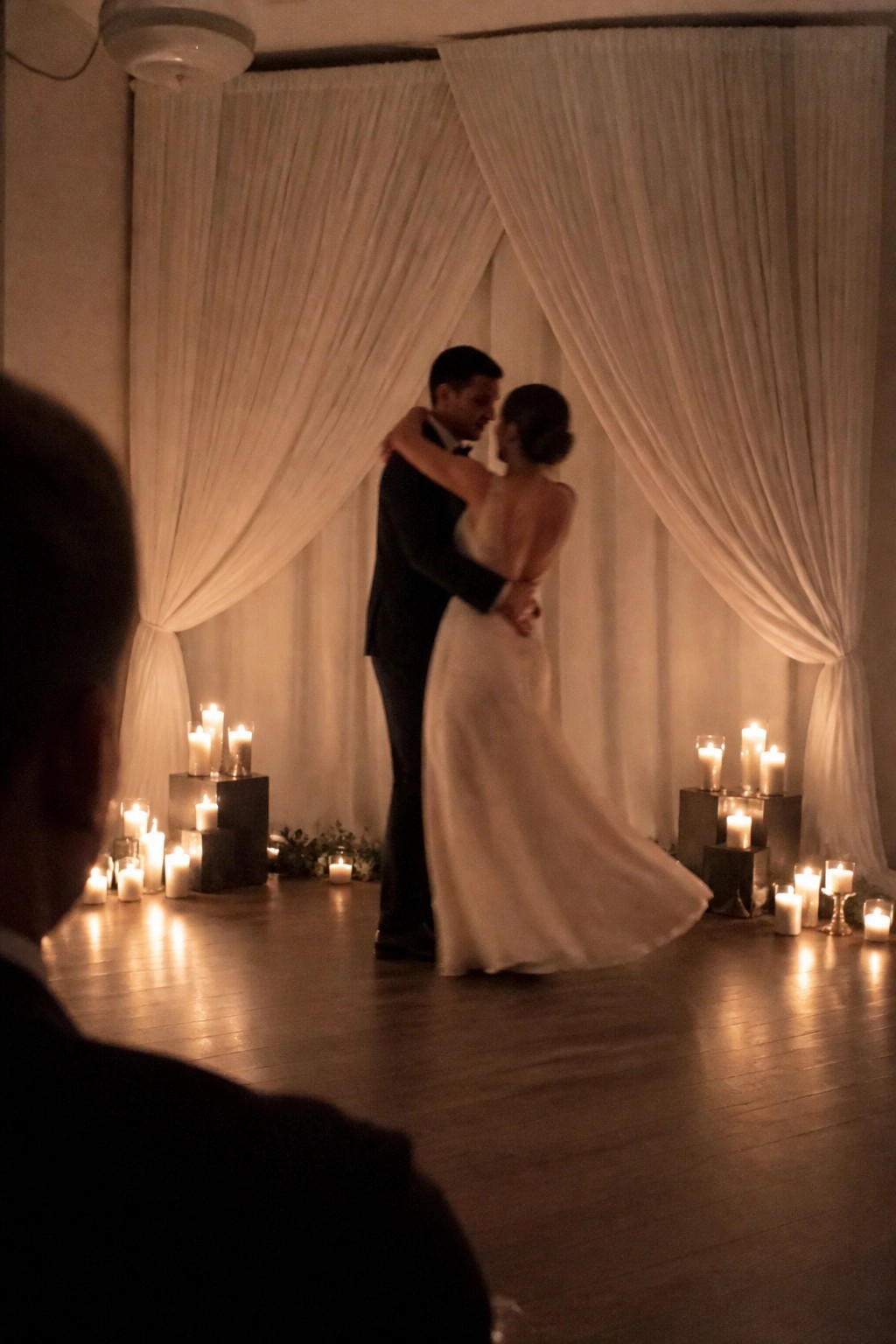 A bride in a white gown and groom in a black tuxedo share a close dance surrounded by glowing candles on blocks with white fabric draping in the background.