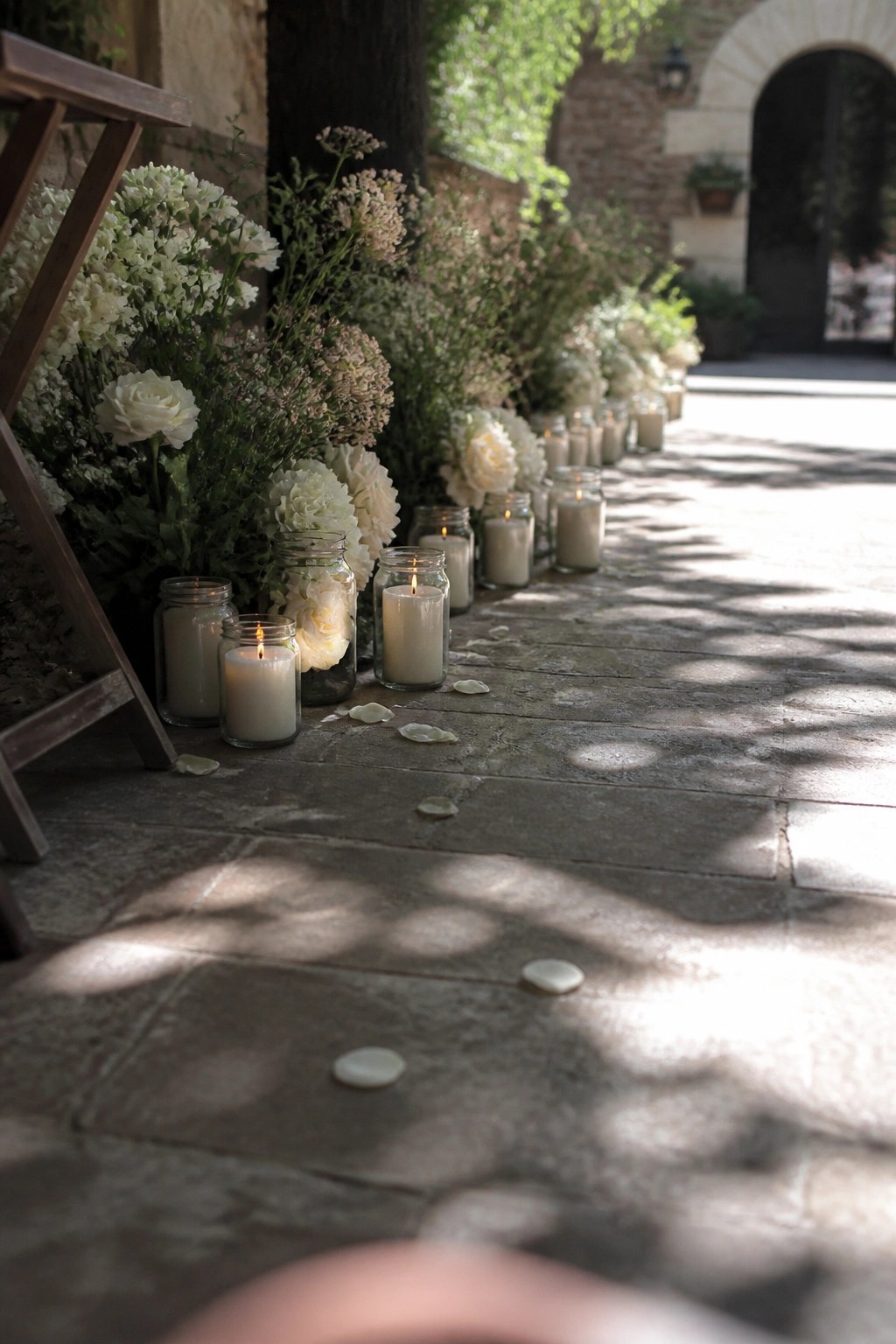 Stone pathway lined with lit white candles in glass jars and clusters of white roses and greenery at an outdoor wedding venue