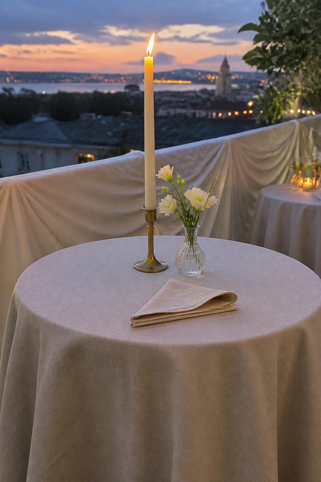 A round table on a balcony at sunset with a tall lit candle in brass, white flowers in glass, folded beige napkin, and draped fabric overlooking a glowing cityscape.