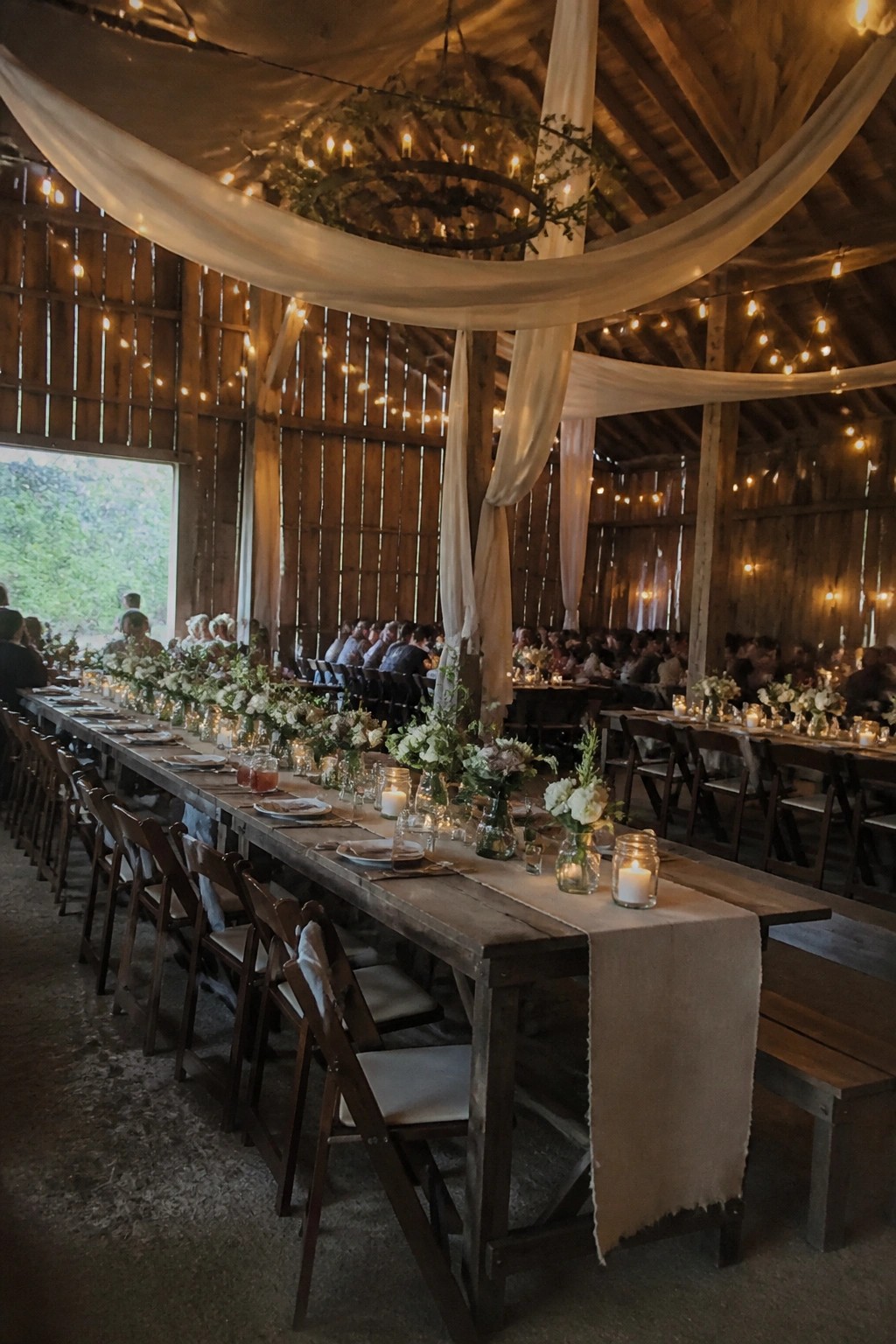 Long wooden tables set with white flowers and glowing candles inside a decorated barn venue with draped fabric and string lights overhead.