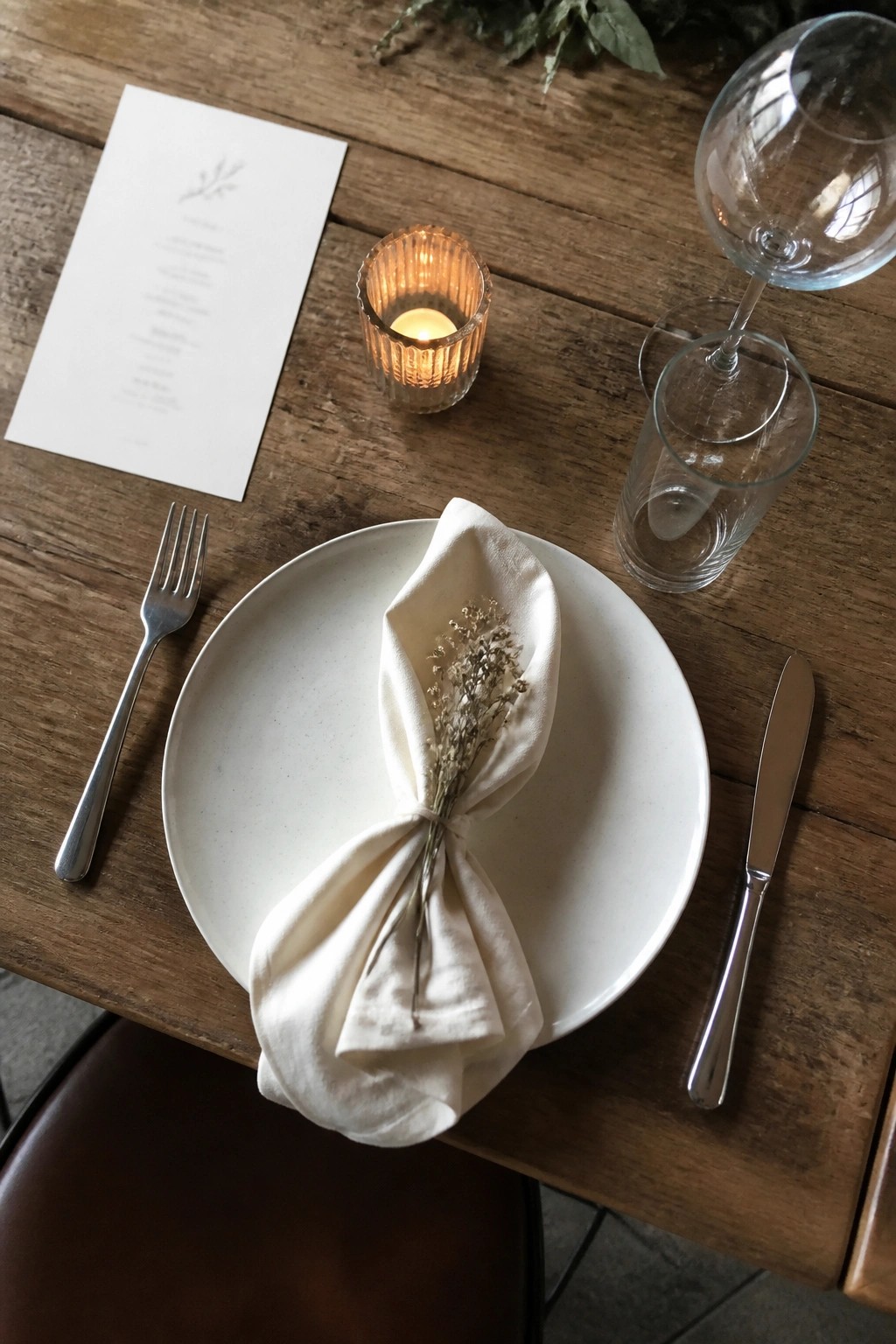 Top down view of a minimalist wedding table setting on rustic wood with a white plate, folded beige napkin tied with dried flowers, silver fork and knife, clear glasses, lit ribbed candle, and illustrated menu card.