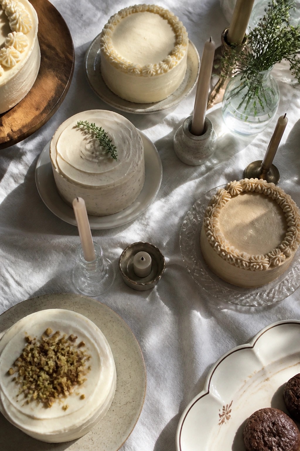 Top down view of multiple small neutral toned cakes arranged with lit and unlit candles in holders on a white tablecloth with soft natural light and greenery accents.