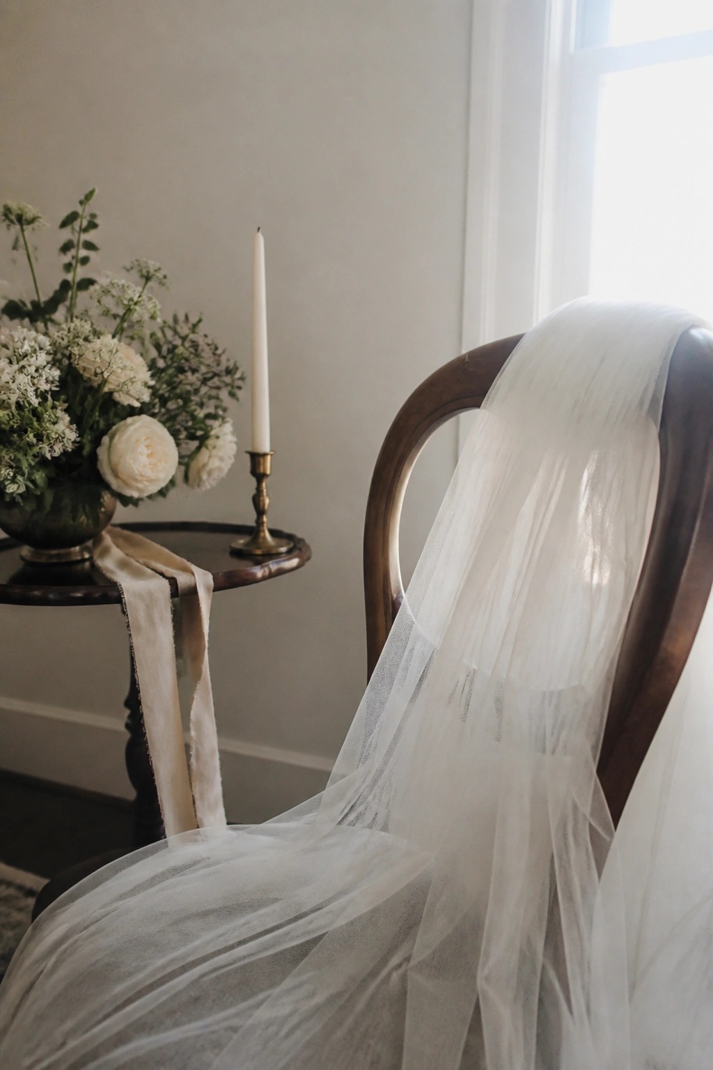 Wooden chair with white tulle draping beside a table featuring white peonies in a vase and a lit candle in a brass holder under soft window light