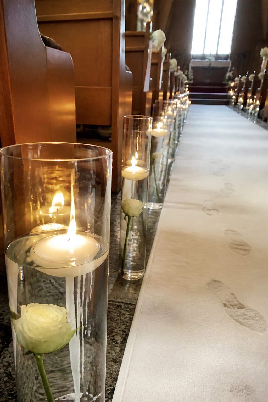 Church wedding aisle lined with tall glass cylinders of floating candles and white roses next to a white runner marked by footprints.