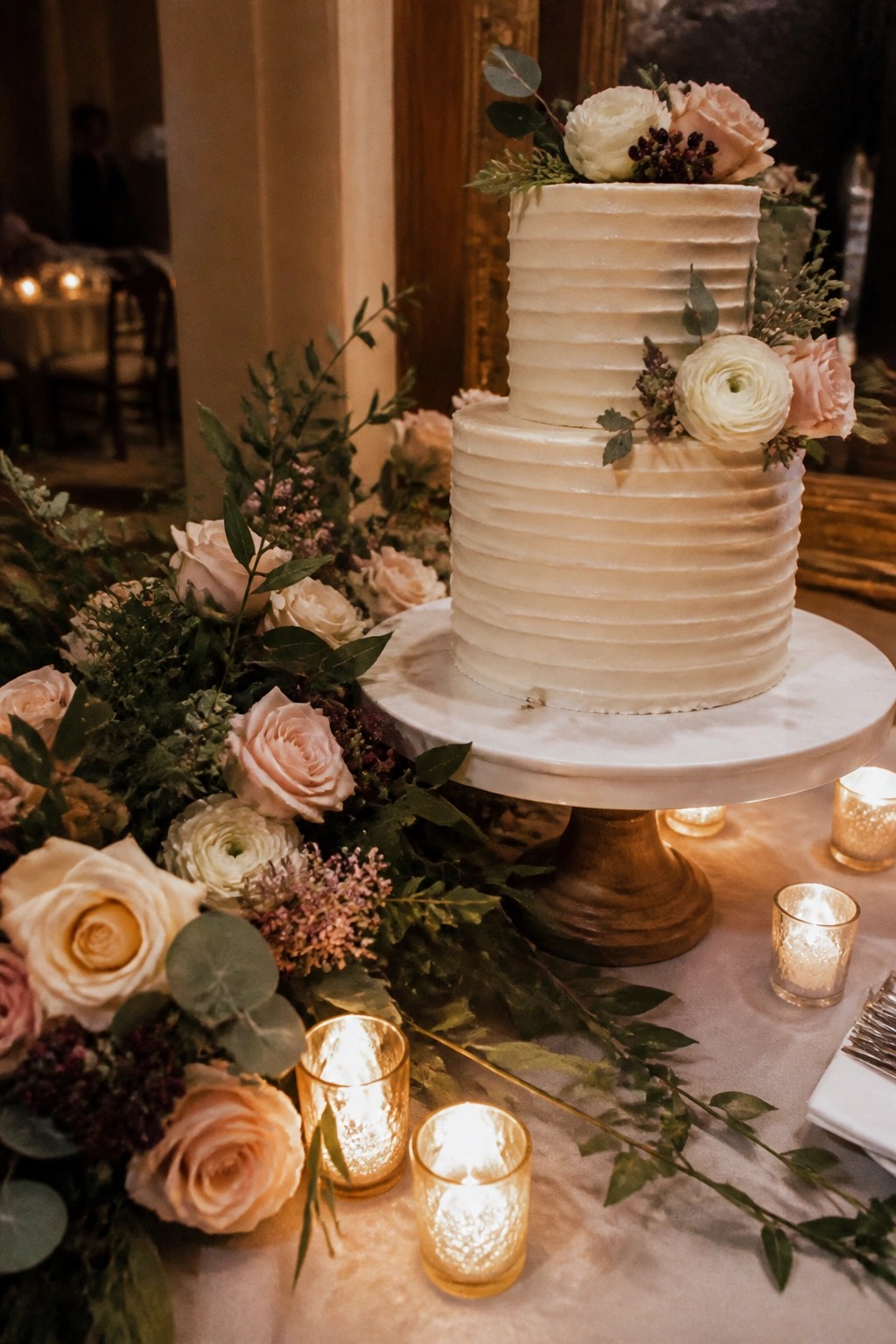 Two tier white wedding cake with blush roses and greenery on marble stand surrounded by floral arrangements and lit candles in glass holders.