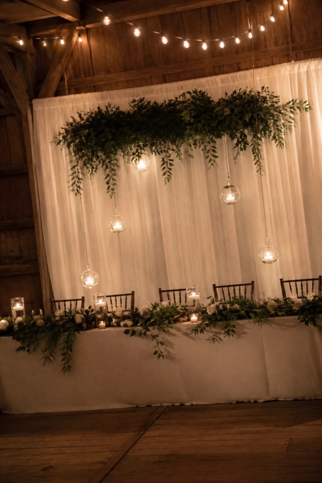 A long wedding head table with white linens and floral garlands sits beneath hanging greenery and glowing candle orbs against a draped backdrop.