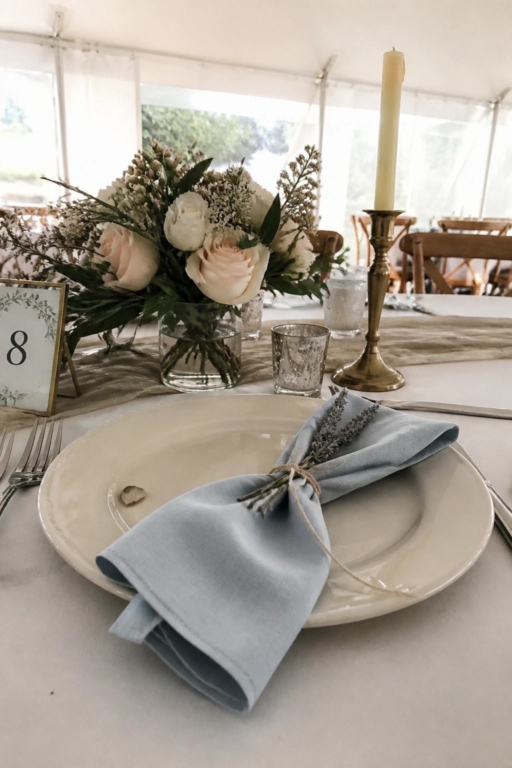 Wedding reception table with white plate, light blue napkin tied with lavender, blush rose centerpiece, brass candle holder, and table number eight in a bright tent.