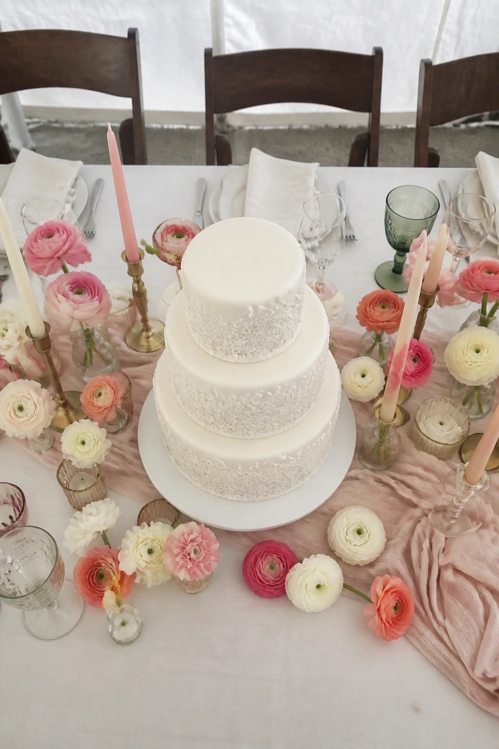 Three tier white wedding cake with lace texture on lower tiers surrounded by pink and white ranunculus flowers and pink candles on a pink runner over a white table.