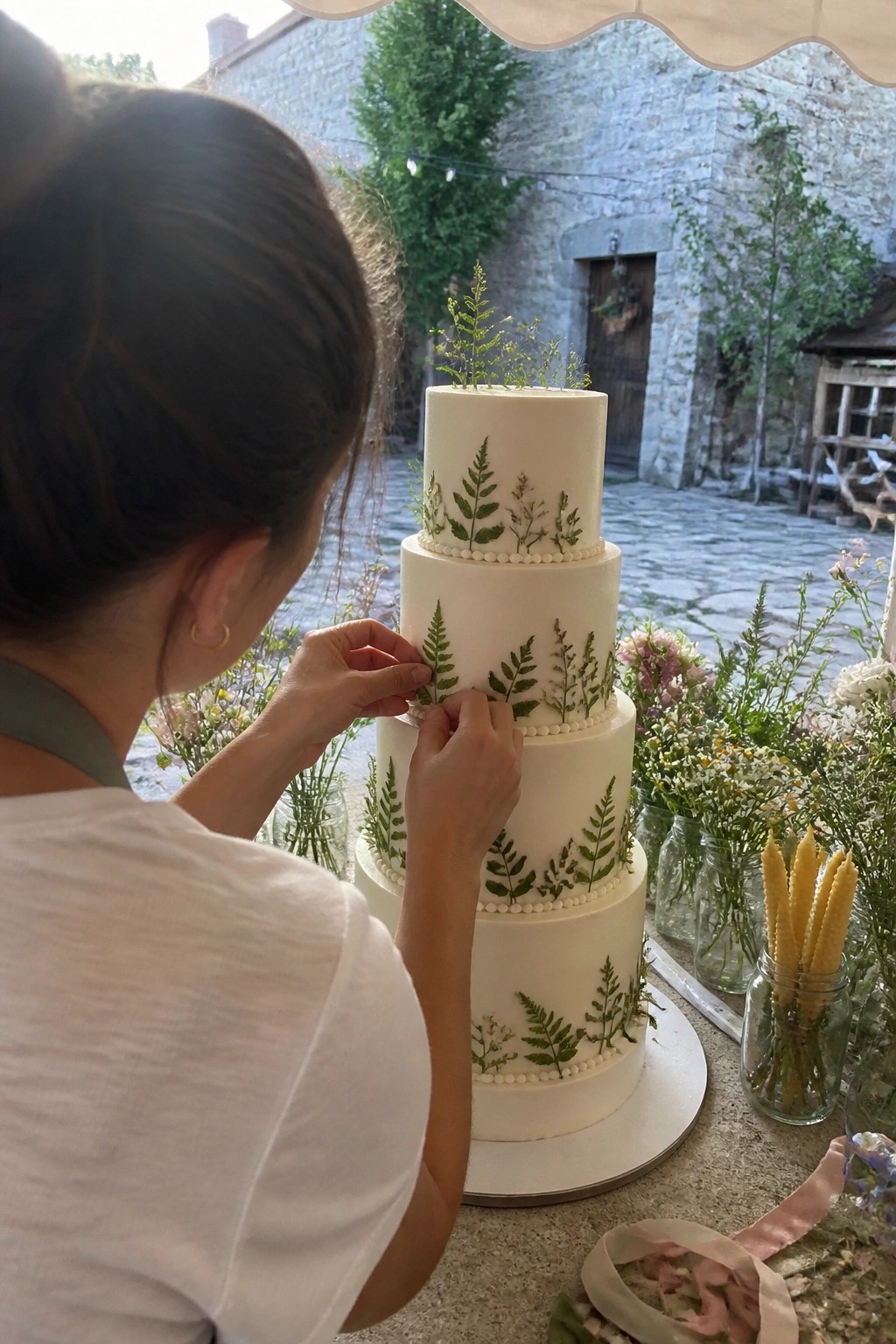 Woman placing green ferns on a four tier white wedding cake decorated with pressed botanicals and pearl borders in an outdoor rustic setting with flowers in jars.
