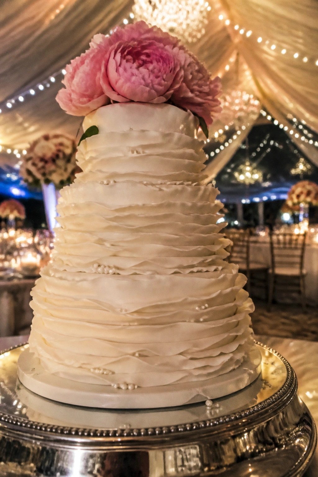 Multi tiered white wedding cake with ruffled texture and pearl accents topped by large pink peonies on a silver stand in an elegant evening venue