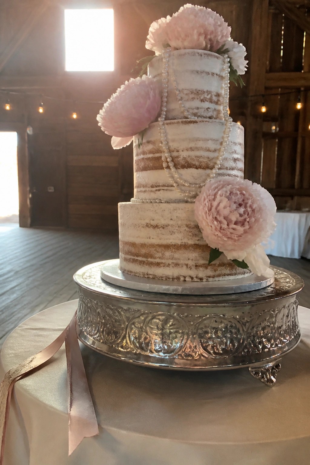 Three tiered semi naked white wedding cake decorated with pink peonies and pearl strands on an ornate silver stand inside a rustic barn venue.