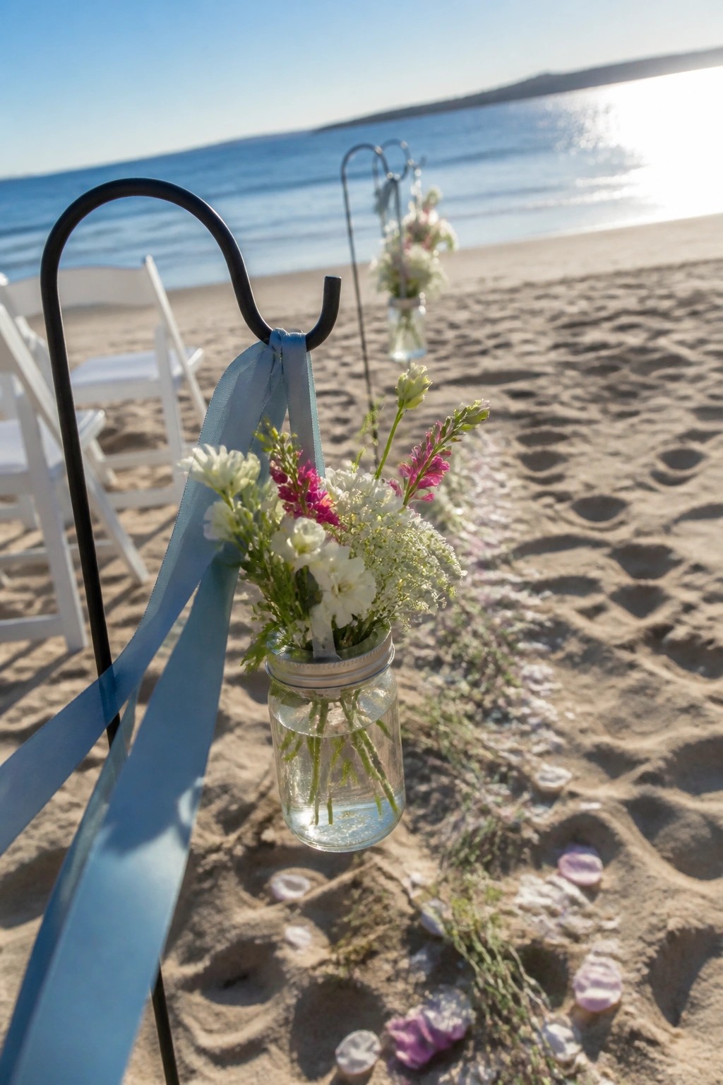 Hanging mason jar with white and pink flowers tied by dusty blue ribbon on a beach wedding aisle with ocean view and scattered petals.
