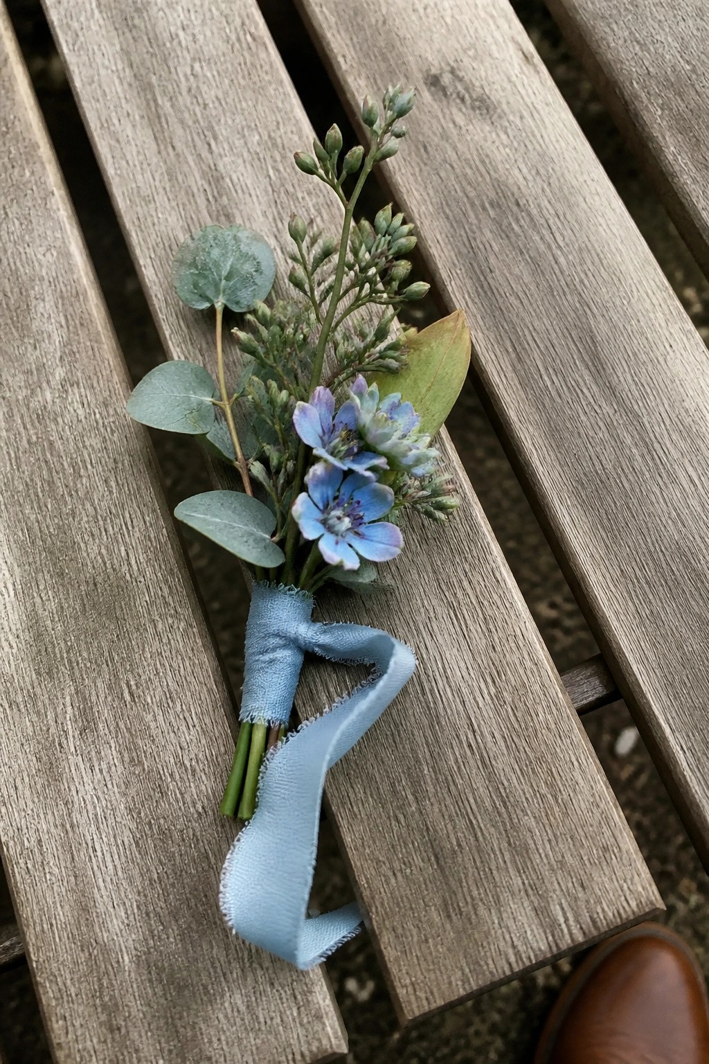 A delicate boutonniere with blue flowers and eucalyptus wrapped in dusty blue ribbon lies on a wooden bench.