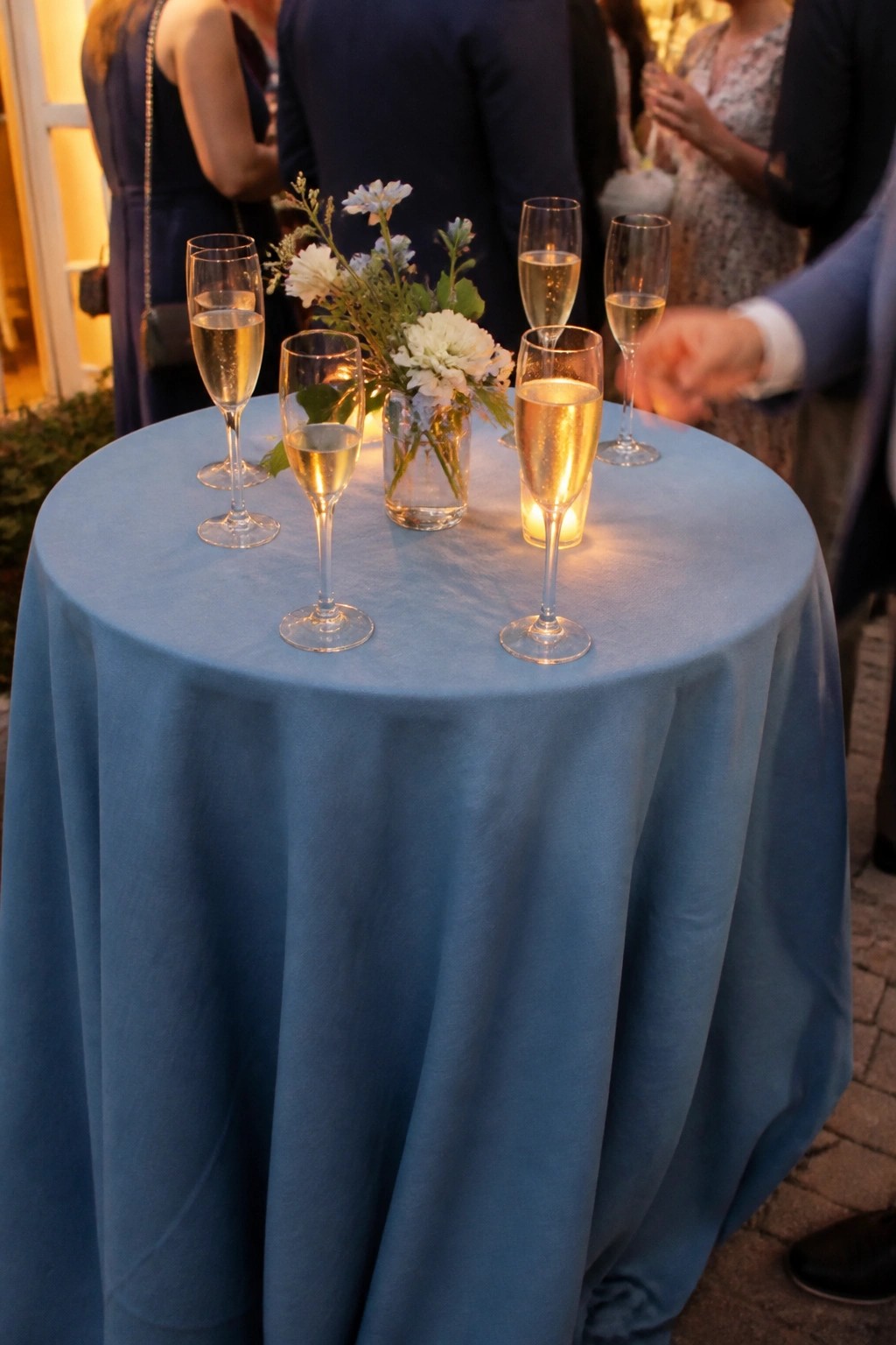 Round table draped in dusty blue linen holds champagne glasses and a small vase of white and light blue flowers with glowing candles at an elegant outdoor wedding reception.