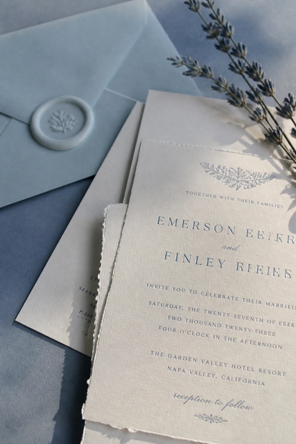 Close up of dusty blue wedding invitation suite with wax seal envelope and lavender sprigs on blue fabric background.