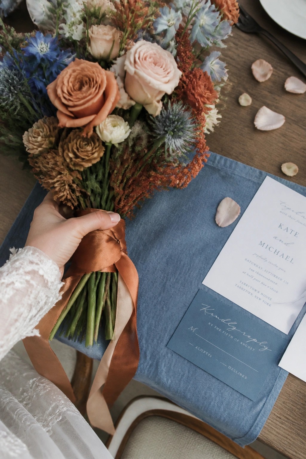 A hand holds a terracotta and peach rose bouquet with blue flowers over a dusty blue linen napkin and wedding invitation on a wooden table with scattered petals.