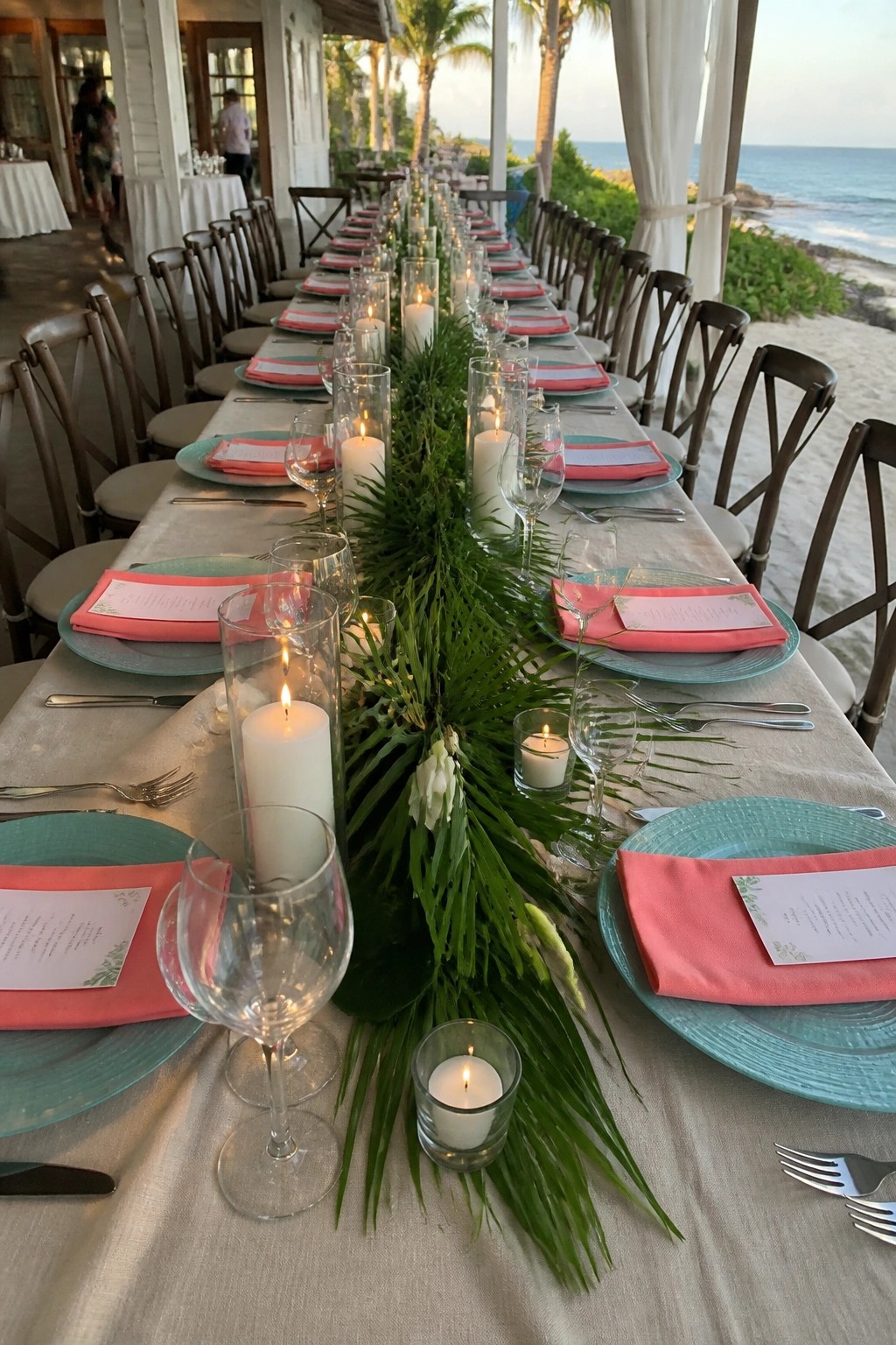 Long beach wedding table with aqua plates coral napkins palm leaf runner and candles overlooking the ocean at dusk