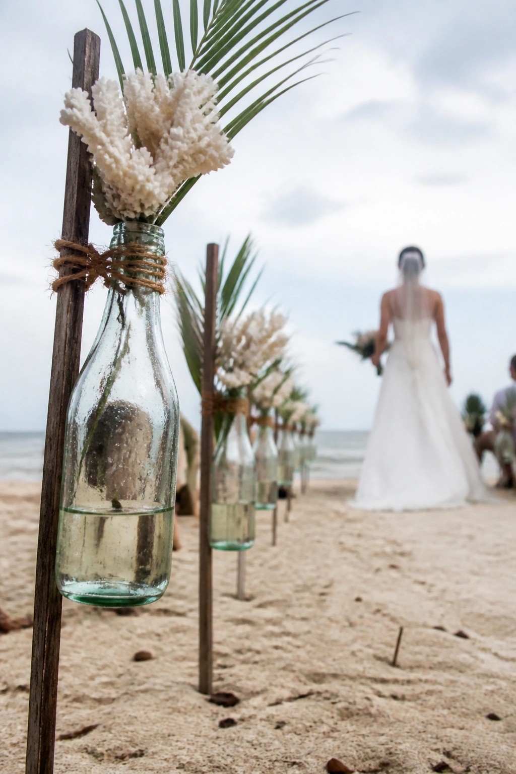 A beach wedding aisle lined with glass bottles on wooden stakes holding white coral flowers and palm leaves with a bride in a white dress walking toward the ocean