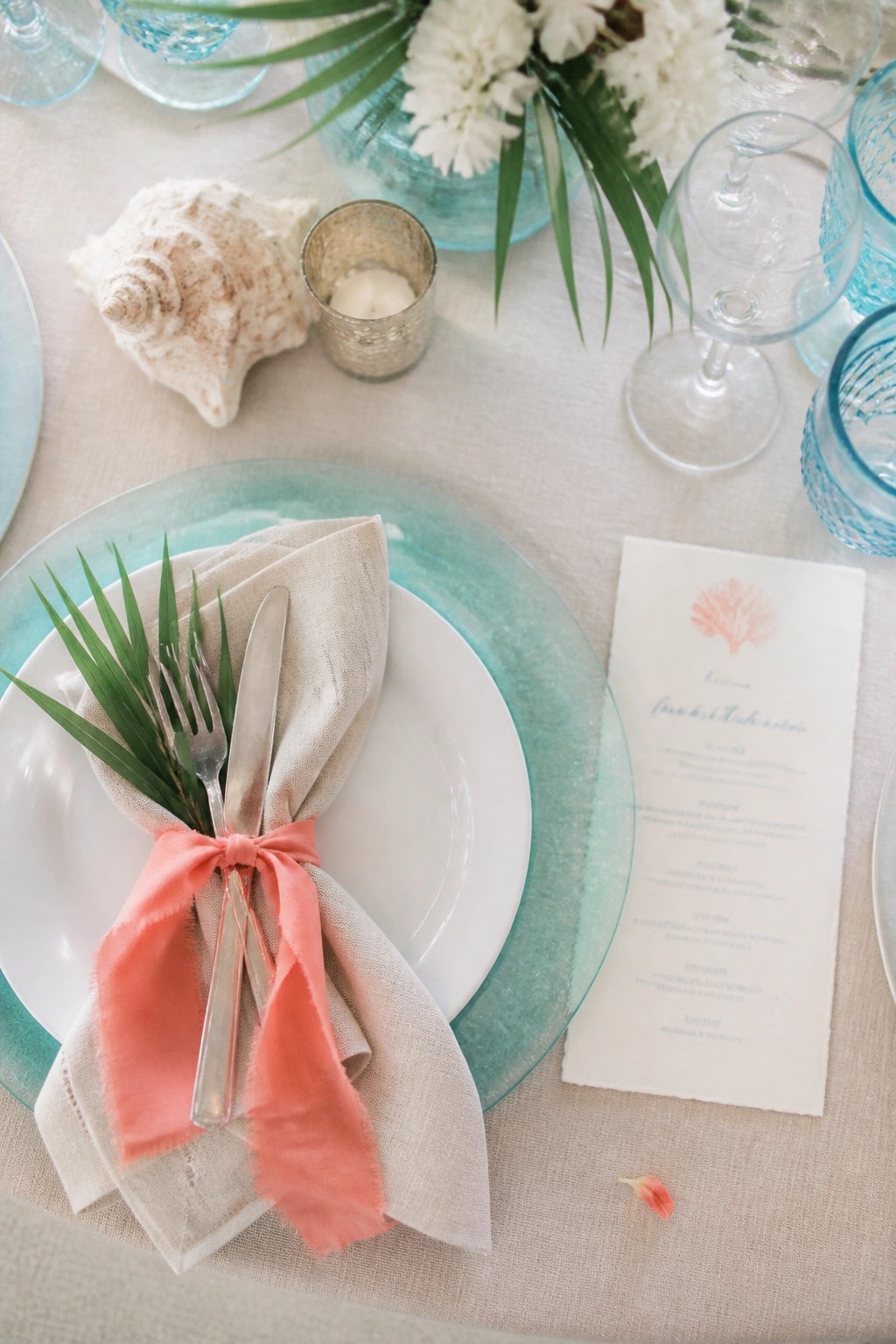 Close up of a tropical beach wedding place setting with white plate on aqua charger, beige napkin tied in coral bow with palm leaves and silverware, seashell, candle, blue glassware, and menu card on sandy tablecloth.