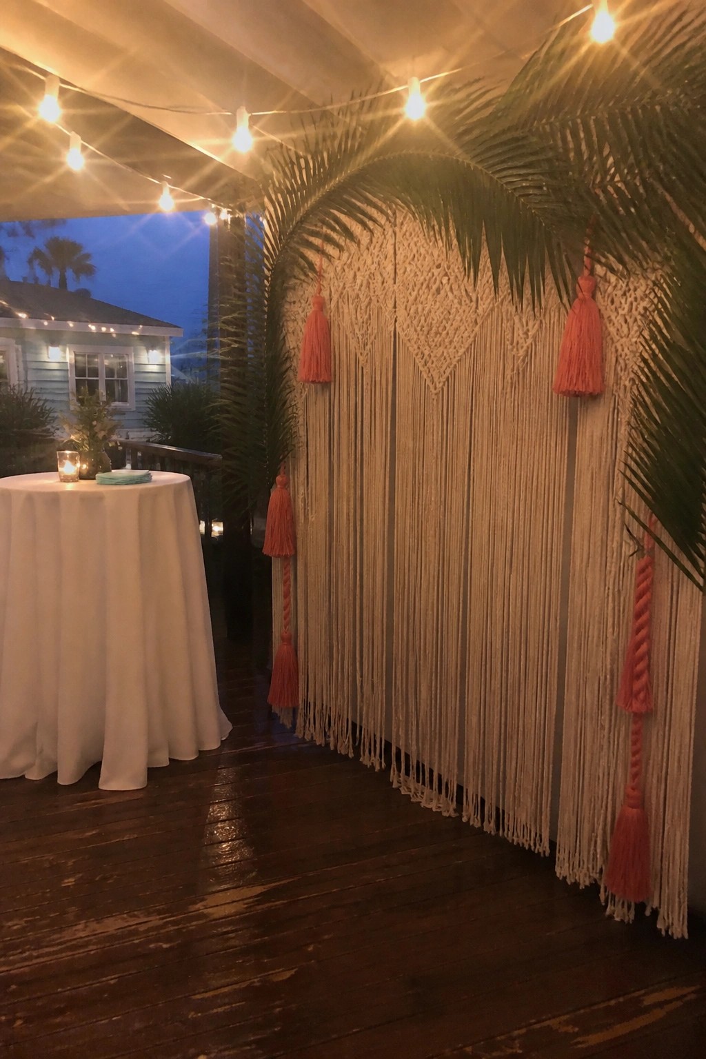 A macrame backdrop with coral tassels and palm leaves on a wooden deck lit by string lights at dusk with a white table nearby