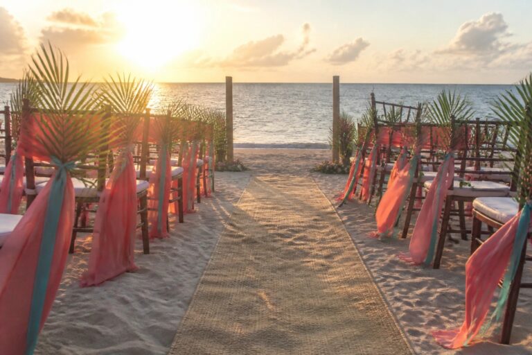 Beach wedding aisle with burlap runner chairs decorated in coral and aqua ribbons palm accents and a white draped arch facing the sunset ocean