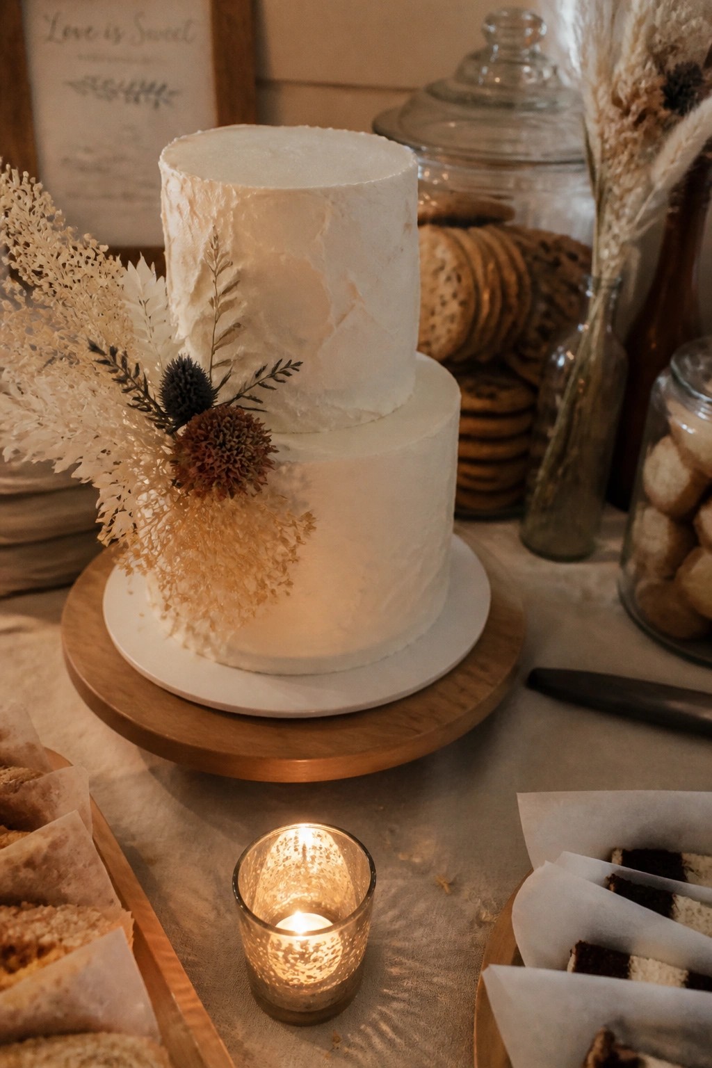 Two tier white wedding cake with dried neutral florals on wooden stand next to glowing candle and assorted cookies in rustic setting