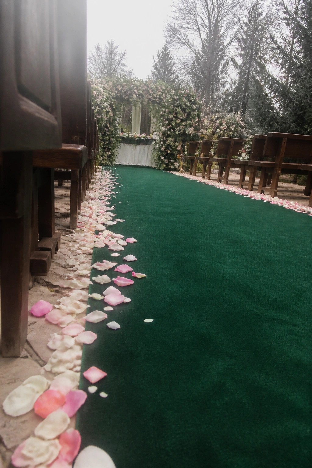 Outdoor wedding aisle with emerald green runner wooden benches pink and white petals and floral arch in misty forest setting