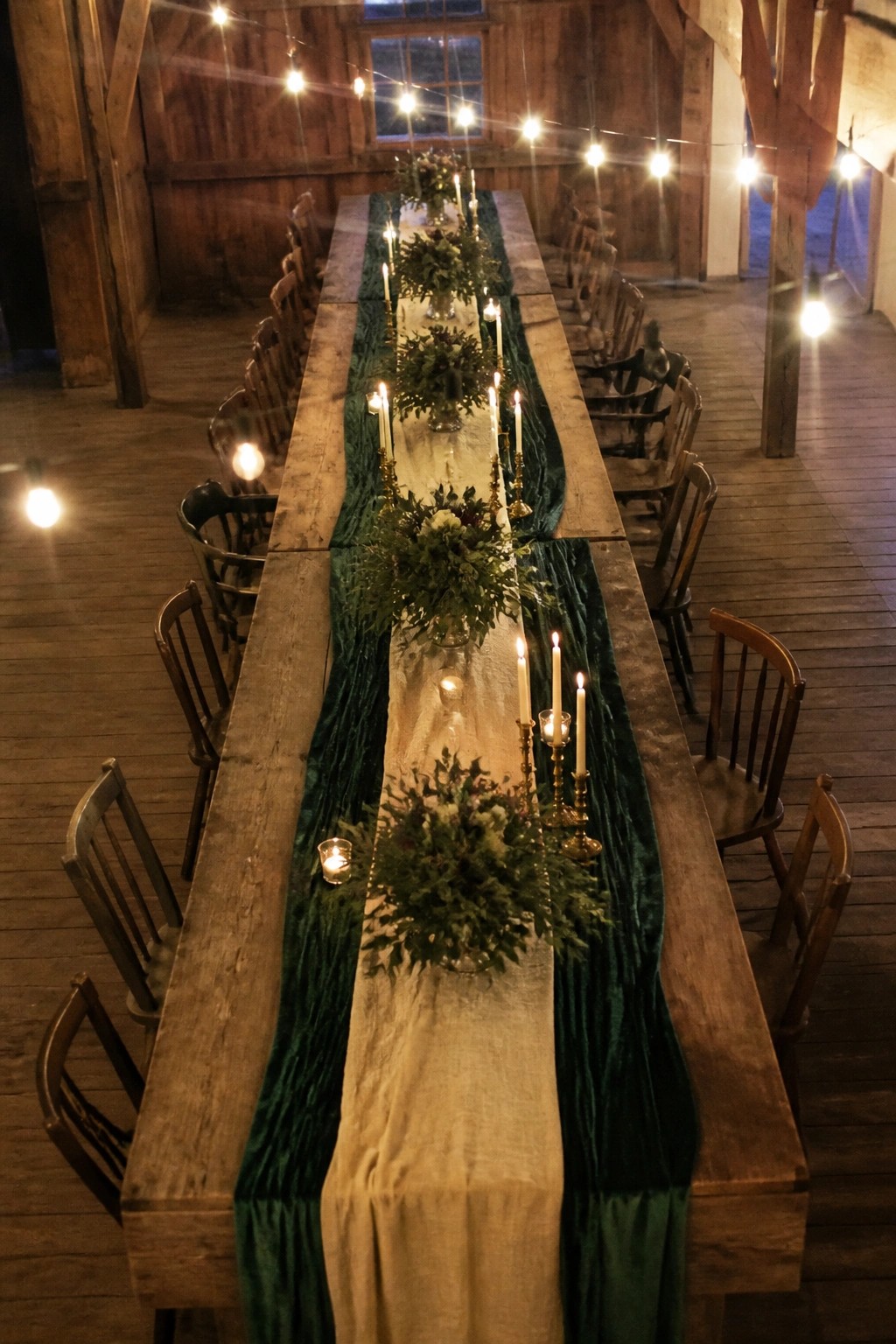 Long rustic wooden wedding table with emerald green velvet runner, green floral centerpieces, white candles in gold holders, and string lights in a barn venue