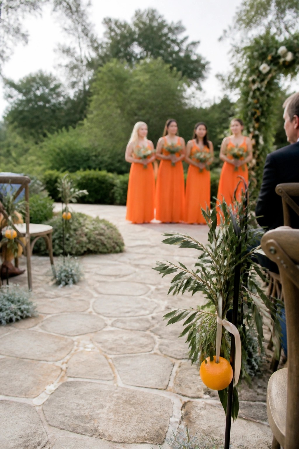 Four bridesmaids in orange dresses stand on a stone patio in a garden with orange decorations on poles and a groom seated nearby.