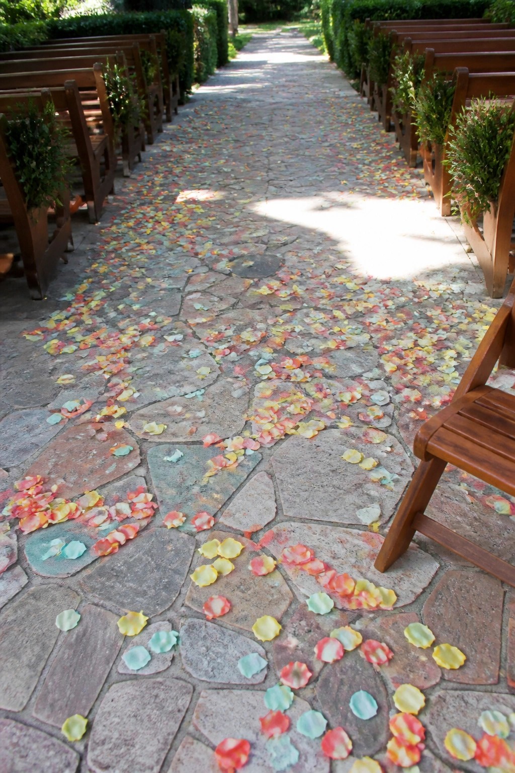 Stone pathway at an outdoor wedding lined with wooden chairs and scattered with bright pink yellow orange and blue flower petals under sunlight
