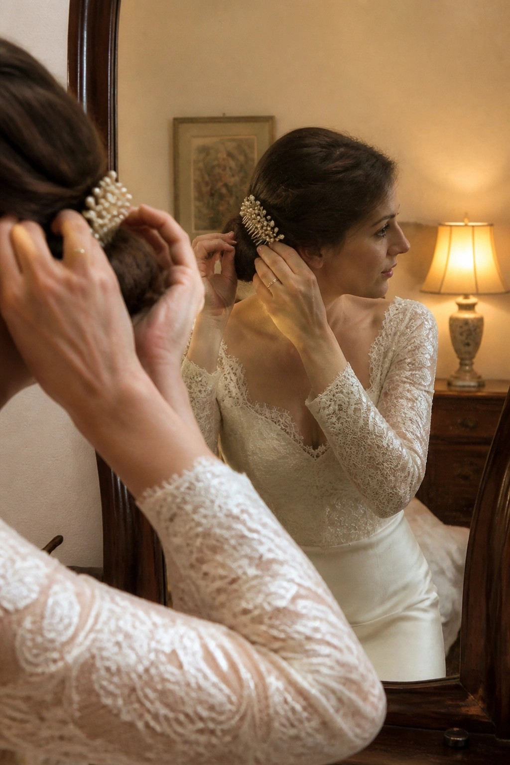 Bride in lace wedding dress adjusts pearl comb in low bun while viewing her reflection in a mirror