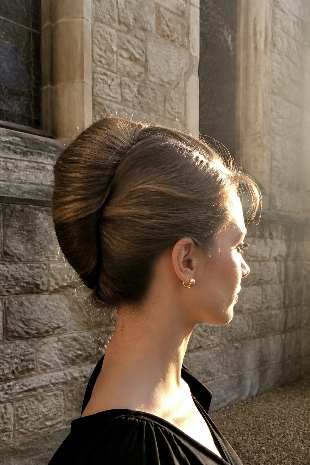 Woman in profile with sleek low bun updo hairstyle against stone wall in sunlight.