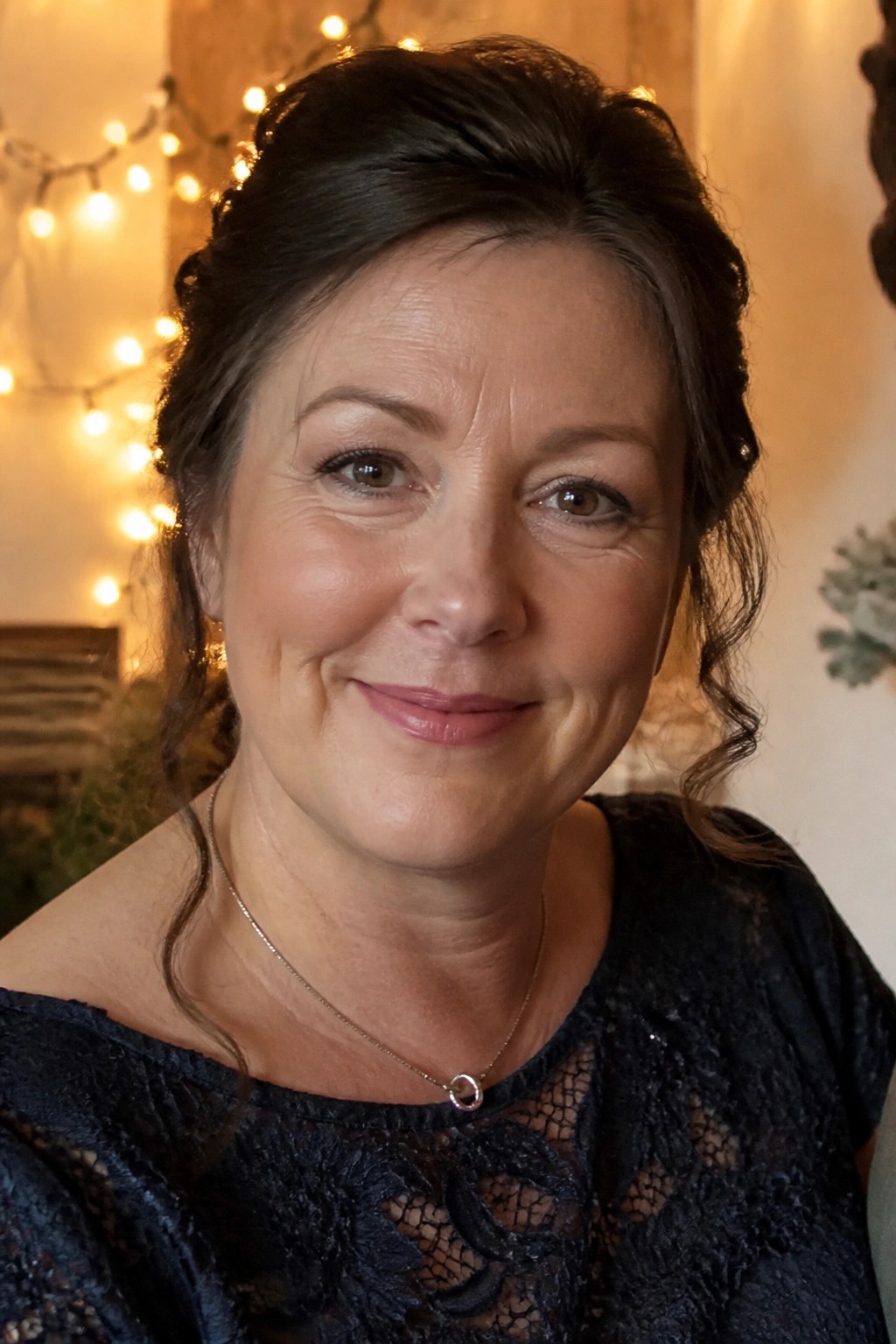 A smiling woman with dark hair in soft waves and a half updo wears a navy lace dress with a necklace against a background of warm string lights.