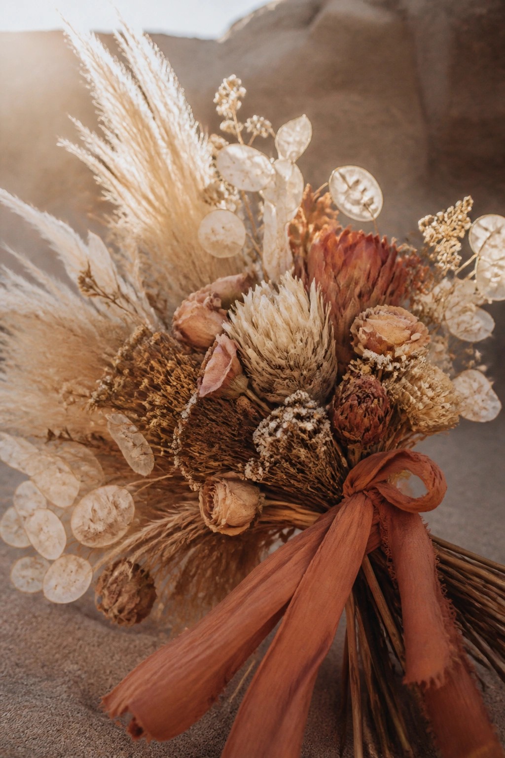 Close up of a dried flower wedding bouquet with pampas grass, proteas, and roses in earthy tones tied with a terracotta ribbon in a desert setting