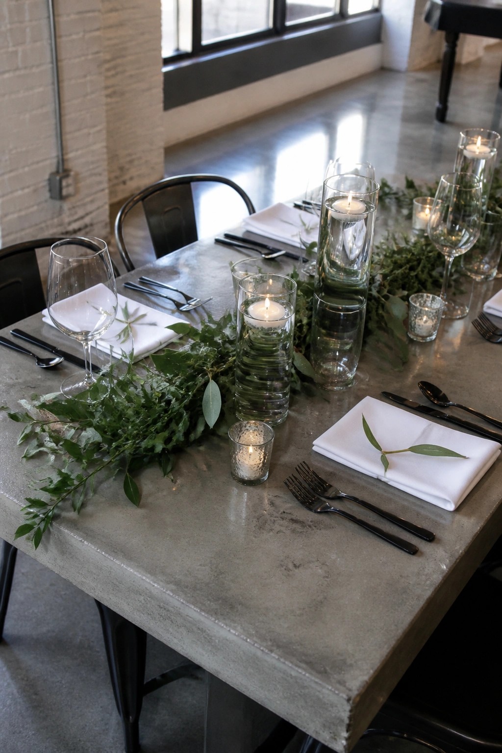 Wedding table on concrete with greenery runner, floating candles in glass cylinders, white napkins with leaves, and black cutlery in an industrial venue with brick walls.
