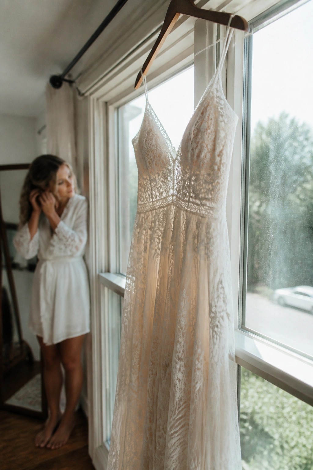 A lace wedding dress hangs by a sunlit window while a bride in a white robe adjusts her hair in the background.
