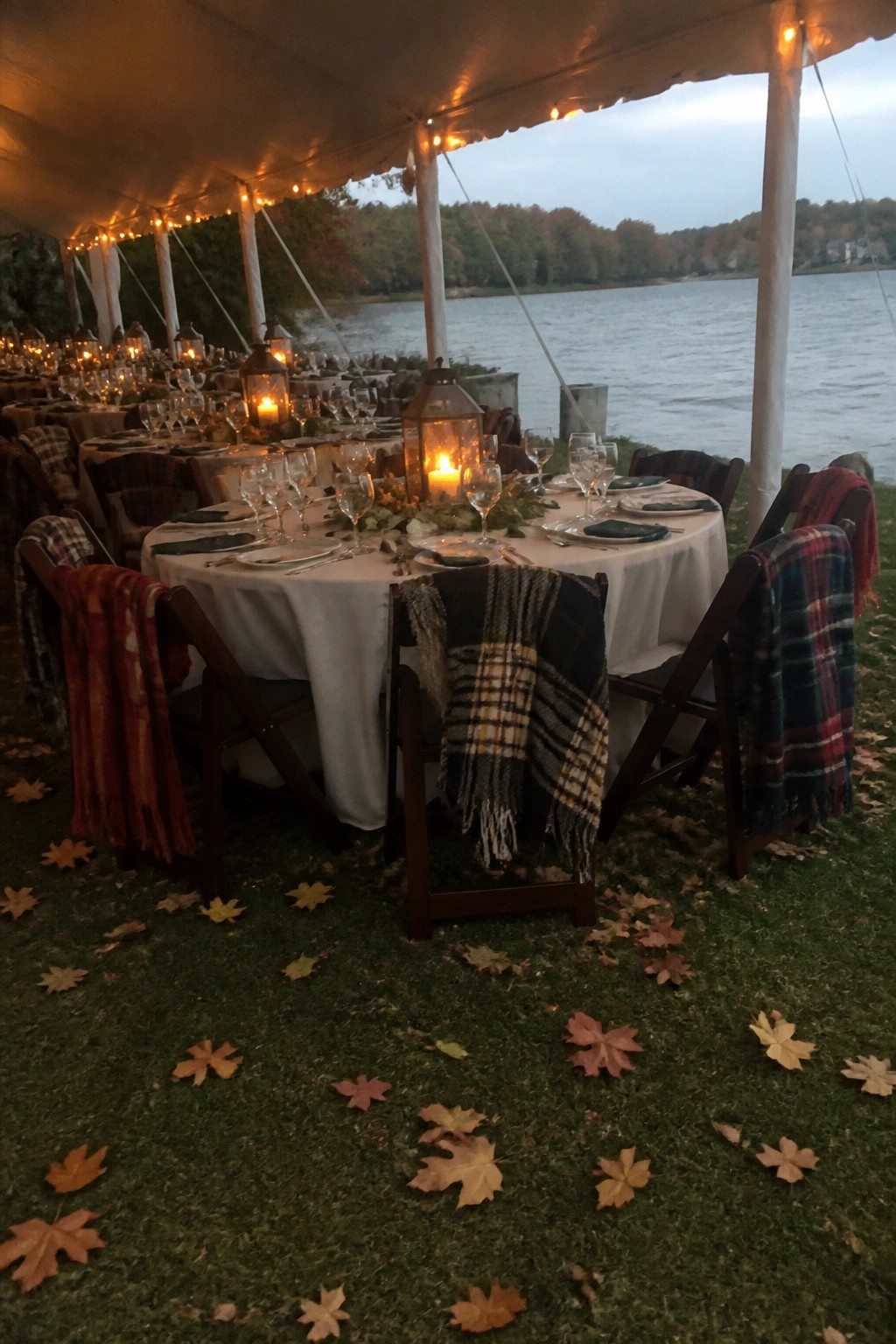 Round dinner table under lit tent by lake at dusk with plaid blankets lanterns and autumn leaves on grass