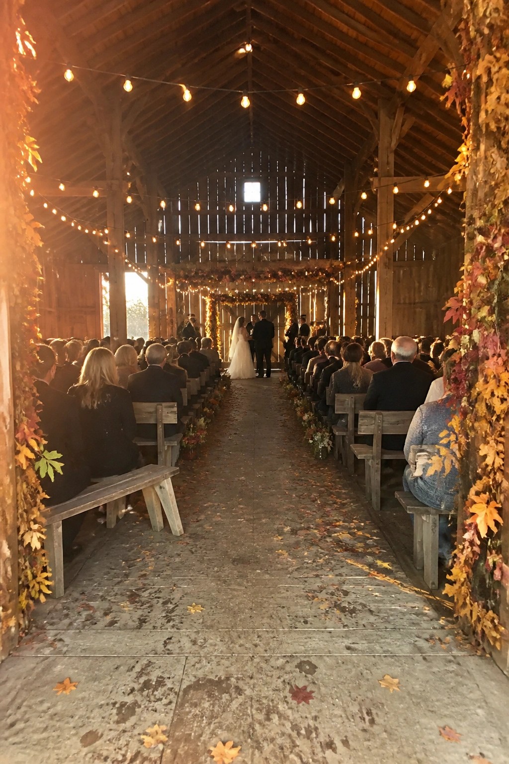 A rustic barn wedding with guests seated on wooden benches, string lights overhead, autumn leaves decorating the space, and a couple at the altar under a floral arch.