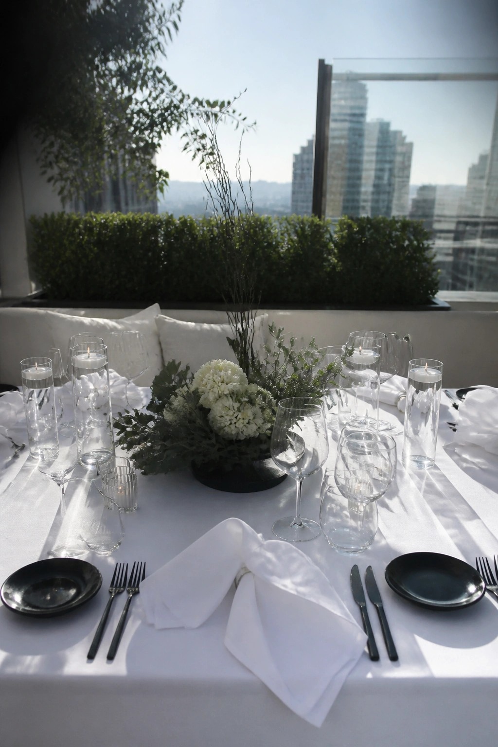 Outdoor wedding table with white linens black dinnerware tall candle holders and white floral centerpiece overlooking city buildings on a sunny day
